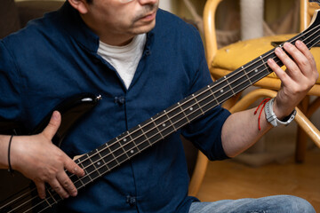 Obraz premium Middle aged man playing black electric bass guitar sitting on floor at home, Musician practicing bass guitar scales while sitting in a living room