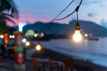 Warm glowing light bulbs hanging along a tropical beach at dusk with a soft bokeh background of mountains, ocean, and evening sky. Moody and serene.
