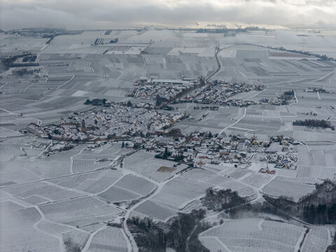 Aerial view of a snow-blanketed village nestled among the geometric patterns of vineyards under a brooding sky, Orschwiller, Grand Est, France.