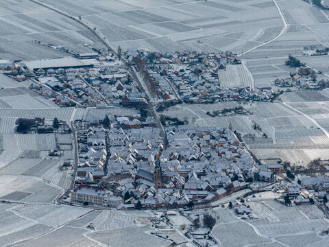 Aerial view of a snow-dusted village nestled amidst vineyard rows, the stark white contrasting with the dark rooftops and leafless vines, Orschwiller, Grand Est, France.