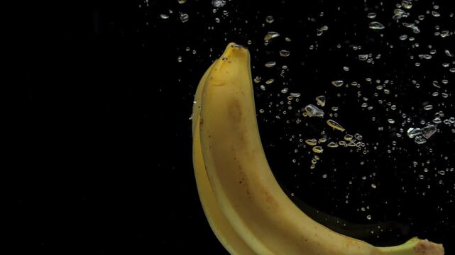 Slow motion bananas falling into transparent water on black background. Fresh fruits splashing in aquarium. Healthy food, diet, air bubbles