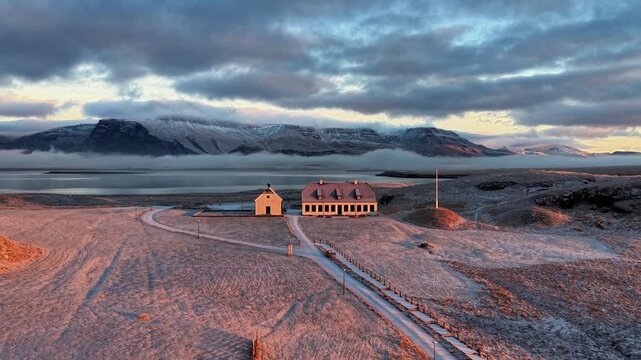Atmospheric cinematic drone footage of a white Scandinavian-style house on a small island in Iceland, illuminated by soft morning sunlight, with calm Atlantic ocean and low clouds covering distant mou