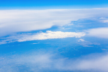 Fototapeta premium Above a patchwork of faint terrain, dense white clouds drift beneath gradient sky that fades from deep blue to pale near the horizon. Aerial view captures serene altitude and atmospheric depth