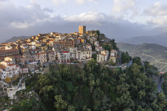 Aerial view of golden sunlight kissing the ancient stone tower atop a hill town amidst lush green foliage, Pollina, Sicilia, Italy.