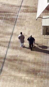 Anciano dependiente con andador y acompa&ntilde;ante trabajador social de compa&ntilde;&iacute;a y ayuda paseando por el patio