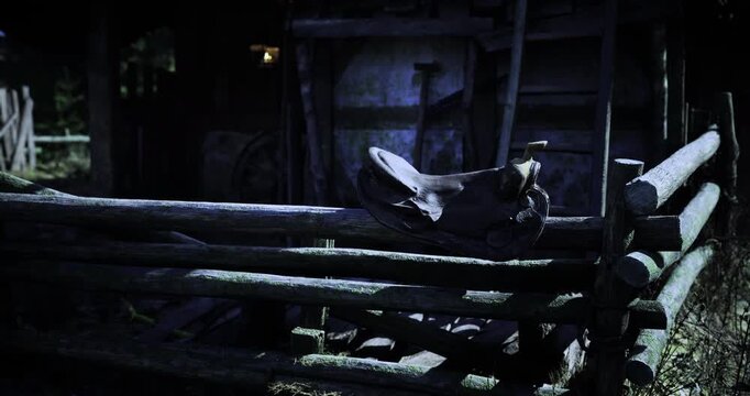 An old saddle rests on a wooden fence surrounding a deserted stable. The setting is dimly lit and evokes a sense of nostalgia in the nighttime atmosphere, presenting a peaceful yet eerie vibe.