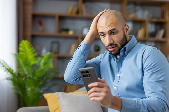 Upset young man in a blue shirt sitting on a sofa at home, looking shocked and frustrated while reading disturbing information on his smartphone with a hand on his head