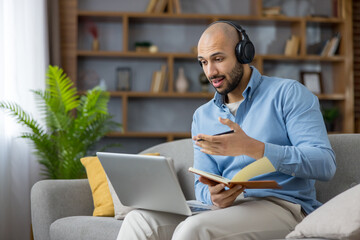 Young man wearing headphones and holding a notebook having a video conference on a laptop from home, actively participating in remote work or online education, communicating and learning
