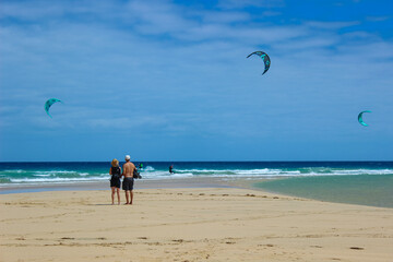 Obraz premium Couple Watching Kitesurfers on Fuerteventura Beach at Sunset