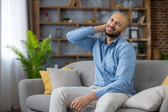 Stressed man sitting on a sofa at home rubbing his neck, suffering from stiff neck and muscle tension after long sedentary work, showing discomfort and fatigue