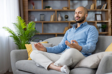 Man sitting cross-legged in lotus on a grey sofa, eyes closed and smiling gently while practicing mindful meditation and finding calm, balance and relaxed inner peace at home