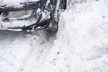 Front wheel and bumper of car pressed tightly into deep snowy rut beside high snowbank, close winter scene showing vehicle stuck in snow, slippery road conditions and difficult cold weather driving.