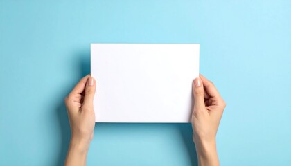 Obraz premium Hands holding blank white paper on light blue background, studio shot.