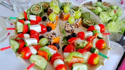 Colorful canapes featuring cheese, tomatoes, cucumbers, olives, grapes, spinach rolls, and pumpkin pieces with sesame seeds arranged on a white plate for festive celebrations