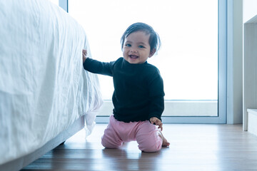A happy Asian baby girl kneeling and smiling joyfully while holding onto the edge of a bed. Process of learning to stand or cruise, symbolizing early childhood development and household milestones.