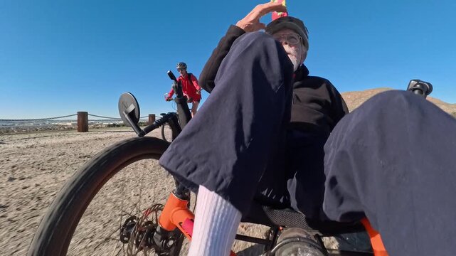 An elderly man rides a recumbent electric bicycle trike across a path on a sunny day with another cyclist ahead of him.