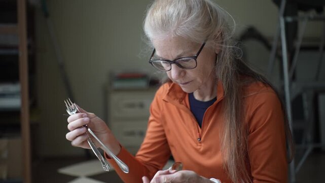 Mature woman looks closely at silver cutlery pieces that hold memories from her family past in her cozy home setting