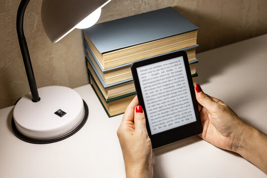 A woman reads an e-reader by the light of a table lamp. A woman reading an e-book at home. reading a book at the table. woman holding e-book. hand holding ereader. e-ink book. 