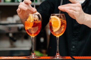 bartender stirs ice with straws in two aperol spritz cocktails on bar counter, orange Venetian cocktail with prosecco lemon and ice cubes