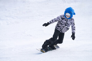 A child wearing a blue helmet skies down a snowy slope on a snowboard. Close-up.