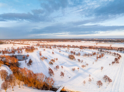 Drone shot shows snow-covered golf course at sunrise with trees, pond and winter sky