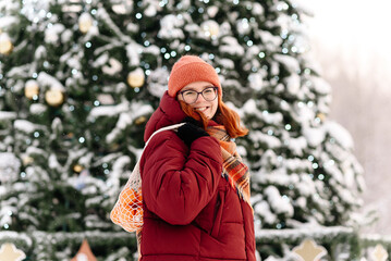 lots of orange tangerines in string bag in young happy woman hands in winter, large christmas tree in bacckground