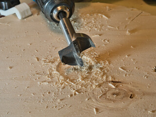 Close-up of Forstner Bit Drilling Wood; High-angle macro shot of a Forstner bit mounted in a power drill, creating a large, precise hole in a light wooden board, surrounded by sawdust.