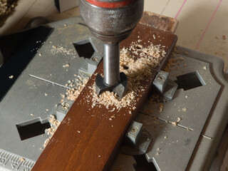 High-angle view of a Forstner drill bit cutting a clean hole into a stained wooden board. Woodworking process in a workshop environment, surrounded by sawdust.