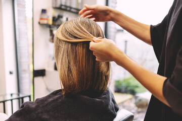 Fototapeta premium Professional hairstylist arranging and smoothing bob hairstyle for female client, back view close up inside modern beauty salon, hand work creating shape