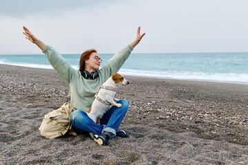 Woman spending time with her Jack Russell Terrier dog in middle season beach. Joyful female hugging...