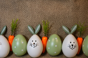 a row of Easter decorations, alternating between white eggs painted with bunny faces and green polka-dotted eggs, interspersed with small artificial carrots against a textured brown fabric background.