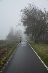 Cold misty day in the Netherlands. Lady walks on Dutch cyclepath in rural countryside heavy fog that limits visibility. Bad weather in nature on footpath