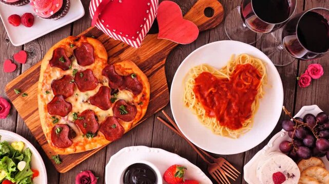 Home cooked Valentines Day dinner. Slow rotating zoom motion. Top view table setting on a dark wood background. Heart shaped pizza, pasta, wine, cheese plate and desserts.