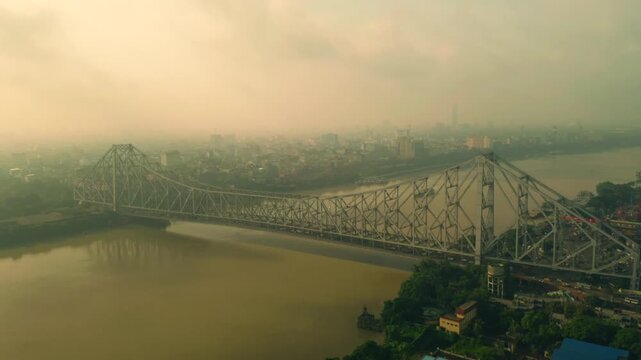 Aerial view of the majestic Howrah Bridge spanning the river, enveloped in a misty haze, connecting the bustling cityscape, Howrah, West Bengal, India.