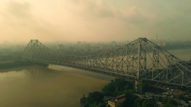 Aerial view of the iconic Howrah Bridge spanning the muddy river, connecting vibrant greenery with the bustling city under a soft, hazy sky, Howrah, West Bengal, India.