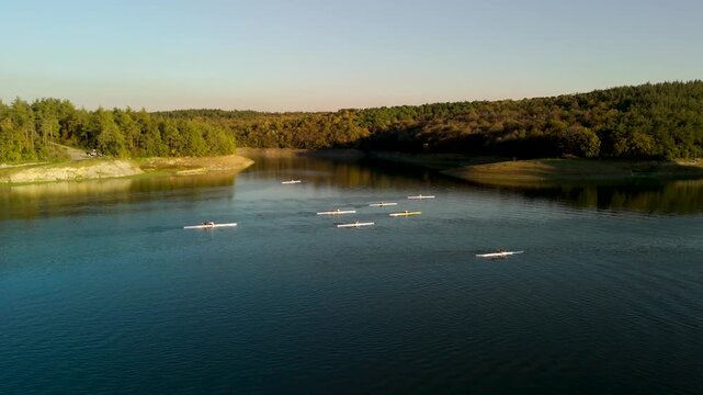 Wide angle view of rowers practicing on a calm lake surface surrounded by lush hills at sunset.