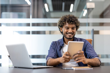 Smiling Indian man sitting at a desk with a laptop and working on a tablet in the office