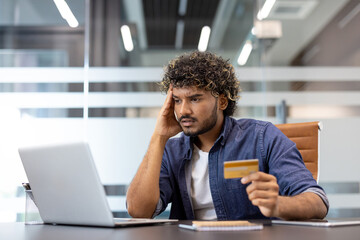Worried young Indian man sitting at a desk in the office, holding a credit card in his hand and looking at the laptop screen in frustration