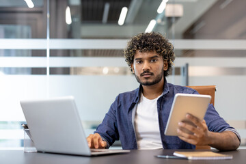 Portrait of a young serious Indian man sitting in the office at a desk with a laptop. holding a tablet in his hand and looking at the camera