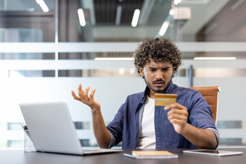 A young Indian man sits at a desk in an office and looks disappointedly at the credit card he holds in his hand