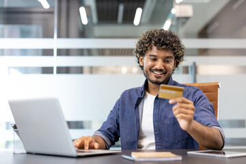 A young Indian man is sitting at a desk in an office, working on a laptop and looking at a credit card he is holding in his hand