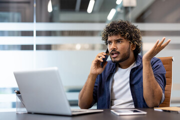 Angry and frustrated young Indian man sitting at a desk in the office and talking emotionally on a mobile phone
