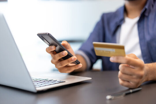 Close-up photo of the hands of a young man sitting at a desk with a laptop, holding a credit card in his hand and using a mobile phone