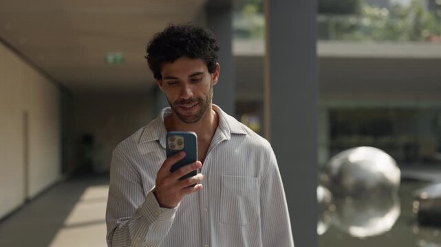 Portrait of curly haired Hispanic man reads message on smartphone as walks on columned passage slow motion. Smiling Latin guy in shirt texts by mobile phone in financial district