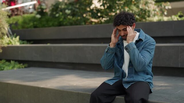 Portrait of tired Latin American man massages temples reducing headache in green park. Hispanic guy deals with migraine attack sitting on parapet in urban district slow motion
