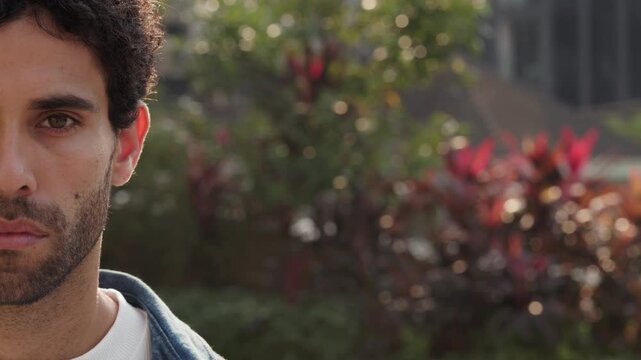 Portrait of Hispanic man face half with beard stands in urban park closeup. Thoughtful Latin American guy with tranquil expression looks at camera on city street. Casual trends