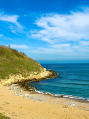 Coastline view with sand and rocks, green hill, and blue sky during daytime at a peaceful beach location