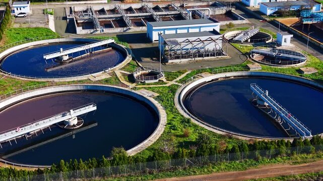 Large sedimentation tanks with mechanical skimmers surrounded by paved roads and green areas. Circular clarifiers with rotating arms. Aerial view.