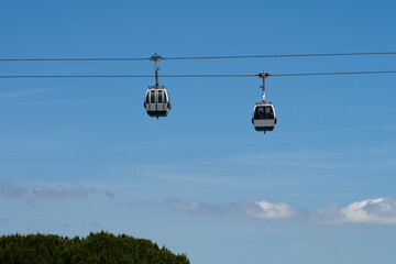 Two cable car gondolas traverse a clear blue sky above a line of green treetops on a sunny day.