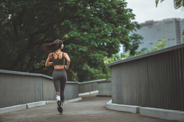 Back view of a young woman athlete in sportswear running and listening to music with headphones on a park bridge with copy space